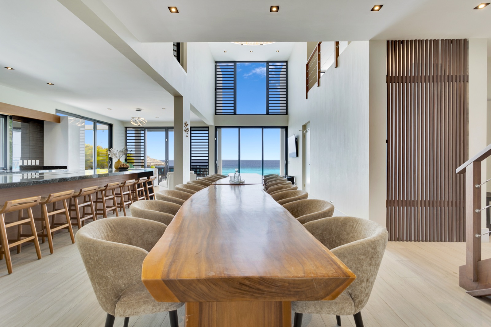 Grand dining room in a luxury Bonaire villa featuring a massive live-edge wood table, double-height ceilings, and panoramic floor-to-ceiling windows with a stunning turquoise ocean view.